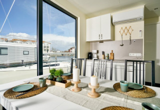 Eettafel op een woonboot met zicht op boten en haven door grote ramen, moderne keuken op de achtergrond.
