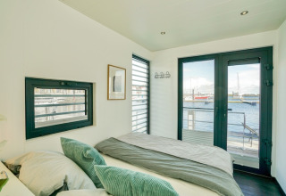 Bedroom on a houseboat with large windows, harbor views, docked boats, and sunlight streaming in.