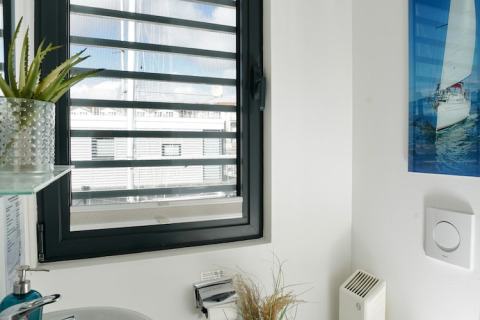 Woonbootbadkamer met groot raam, groene plant, zeilbootfoto en moderne witte muren en inrichting.