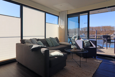 Moderne woonkamer op een woonboot in Duitsland met grote ramen, zetel en uitzicht op de haven.