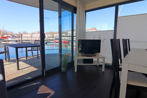 Soggiorno su una casa galleggiante con tavolo da pranzo, televisore e terrazza vista porto turistico.
