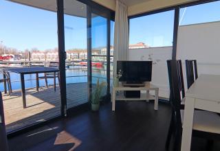 Soggiorno su una casa galleggiante con tavolo da pranzo, televisore e terrazza vista porto turistico.