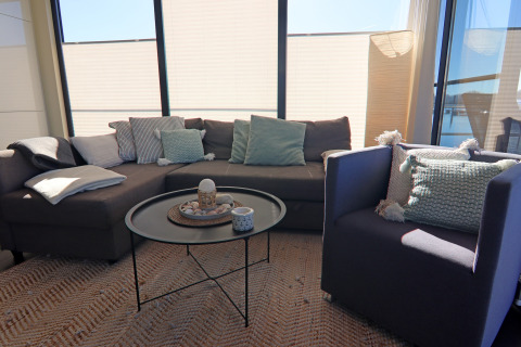 Salon moderne sur une péniche avec grandes fenêtres, canapés gris, coussins décoratifs et table ronde.