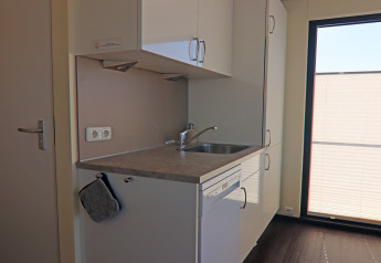 Compact kitchenette with sink, dishwasher and large window on a houseboat, illuminated by sunlight.