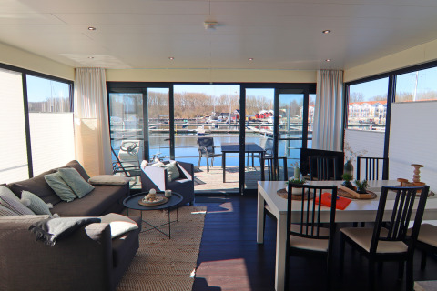 Lichte woon- en eetruimte op een woonboot met grote ramen en zicht op de jachthaven en het water.