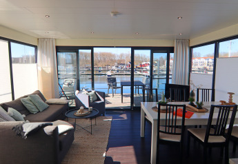 Lichte woon- en eetruimte op een woonboot met grote ramen en zicht op de jachthaven en het water.