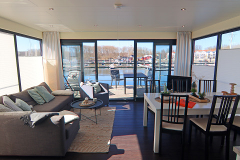 Salón de casa flotante con sofá, mesa de comedor y grandes ventanales con vista al puerto exterior.