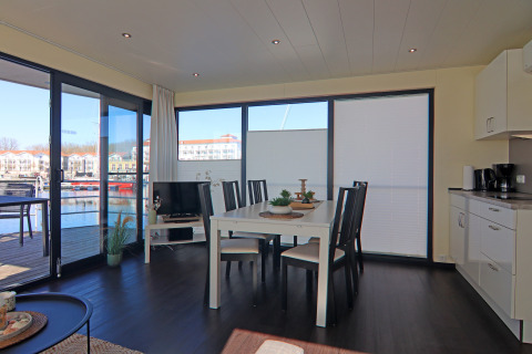 Interior luminoso de una casa flotante con comedor, cocina y vistas al puerto deportivo desde las ventanas grandes.