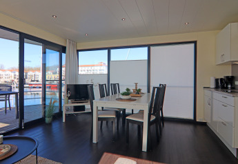 Interior luminoso de una casa flotante con comedor, cocina y vistas al puerto deportivo desde las ventanas grandes.
