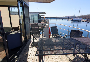 Zicht vanaf het terras van een woonboot met tuinset, stoelen, reddingsboei en boten op de achtergrond.