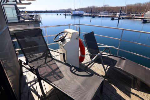 Terrasse ensoleillée d'une péniche avec chaises longues, volant et vue sur le port et les bateaux.
