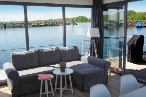 Soggiorno su una casa galleggiante con divano grigio, grandi finestre, vista sull'acqua e terrazza.