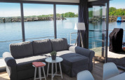 Woonkamer op een woonboot met grijze bank, grote ramen, uitzicht op water en terras met groen buiten.