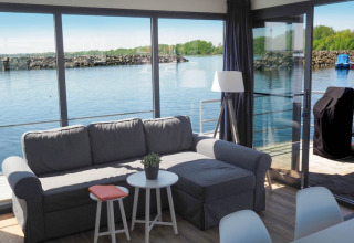 Soggiorno su una casa galleggiante con divano grigio, grandi finestre, vista sull'acqua e terrazza.