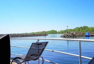 Vista da una houseboat con sedia, ringhiera, acqua calma, moli di pietra e alberi sotto un cielo azzurro.