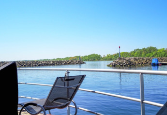 View from a houseboat featuring a chair, railing, calm water, stone barriers, and trees under blue sky.