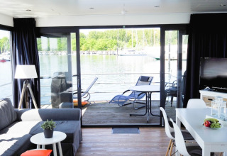 Interno moderno di una houseboat con salotto, tavolo da pranzo e vista sul ponte e sull’acqua dalle vetrate.