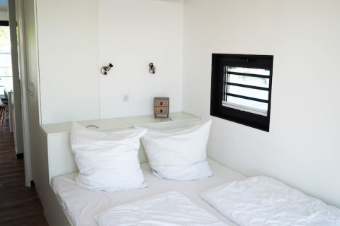 Zona de descanso en una casa flotante con almohadas blancas, lámparas modernas y una pequeña ventana.