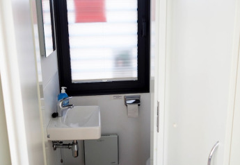 Petite salle de bain sur une péniche avec lavabo blanc, distributeur de savon bleu, fenêtre et papier toilette.