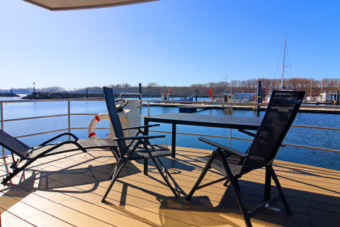 Sunny view from a houseboat deck with chairs and table, overlooking a marina and boats in the background.