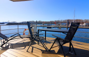 Vista soleada desde la cubierta de una casa flotante con sillas y mesa, y un puerto deportivo al fondo.