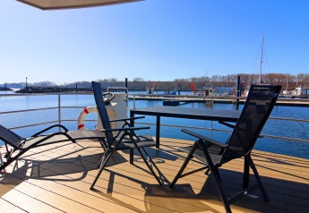 Vue ensoleillée depuis le pont d'une péniche avec chaises et table, et un port de plaisance en arrière-plan.