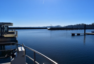 Vue depuis une péniche sur un port calme, avec un quai, ciel bleu et arbres à l’horizon en plein soleil.