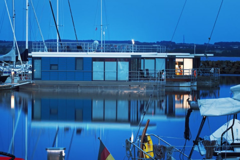 A modern houseboat illuminated at dusk, docked in a marina with its reflection on calm blue water.