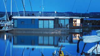 Een moderne woonboot met verlichting bij valavond, aangemeerd in een haven en weerspiegeld op het water.
