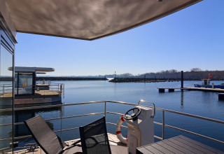 View from a houseboat deck with chairs and steering wheel, overlooking calm waters and a marina on a clear day.