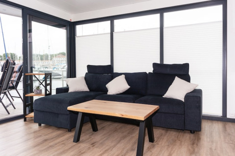 Modern living area on houseboat with dark sofa, wood table, and large windows overlooking marina.