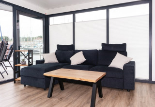 Salón moderno en una casa flotante con sofá oscuro, mesa de madera y grandes ventanas al puerto.