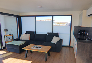 Modern living area on a houseboat featuring a large sofa, coffee table, kitchenette, and big windows.