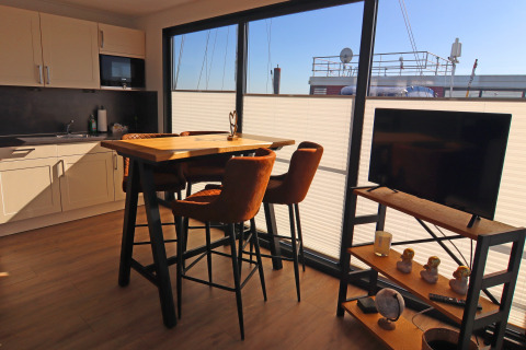 Modern houseboat interior with dining table, chairs, kitchenette, TV, and large windows to the deck.