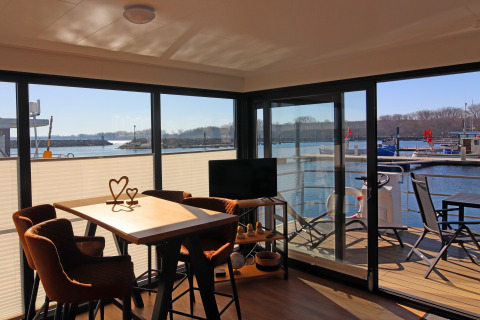 Woonbootinterieur met een zonnige eethoek, tv en terras met stoelen, zicht op de jachthaven.