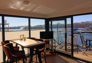 Houseboat interior with a sunny dining area, TV, and outdoor deck with chairs overlooking a marina.