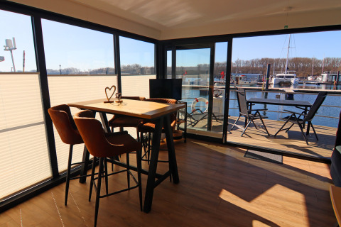 Interior of a houseboat showing dining area with table and chairs, deck and marina visible outside.