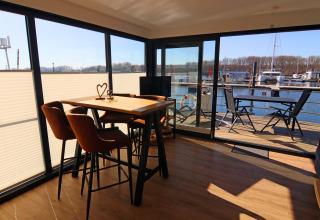 Interior of a houseboat showing dining area with table and chairs, deck and marina visible outside.