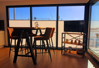 Sunny houseboat interior with a dining table, chairs, TV, and large windows overlooking the water.