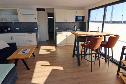 Modern houseboat kitchen and living area with large windows, dining table, and brown chairs.