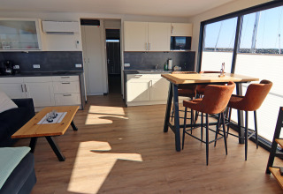 Modern houseboat kitchen and living area with large windows, dining table, and brown chairs.