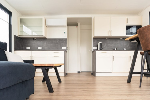 Modern houseboat interior with a sofa, kitchen with white cabinets, wooden floor, and bar stool in Germany.