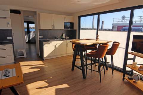 Sunny houseboat interior with large window, dining table, four chairs, and a modern kitchen.