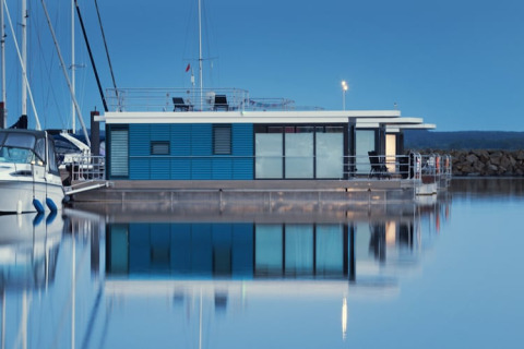 A modern houseboat with a blue exterior and large windows floats peacefully in a calm marina at dusk.