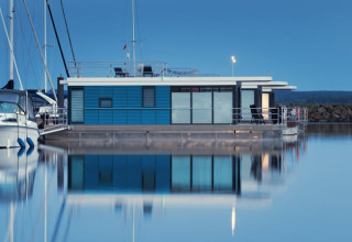 A modern houseboat with a blue exterior and large windows floats peacefully in a calm marina at dusk.