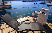 Solbeskinnet terrasse på en husbåd med liggestol, redningskrans og udsigt til roligt havnevand.