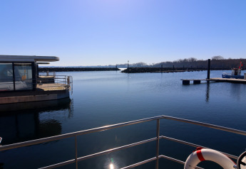 Zicht vanaf een woonboot op een rustige haven met blauwe hemel en kabbelend water in de zon.