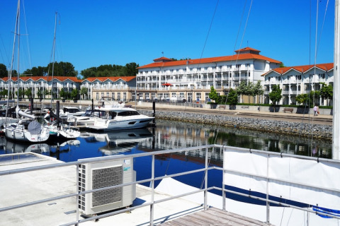 Houseboat docked in a peaceful marina with yachts, waterfront walkway, and red-roofed buildings behind.