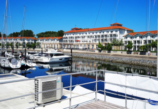 Houseboat docked in a peaceful marina with yachts, waterfront walkway, and red-roofed buildings behind.