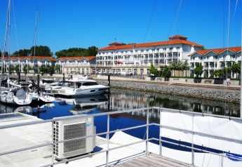 Casa flotante atracada en un puerto tranquilo con yates y edificios de tejados rojos al fondo.